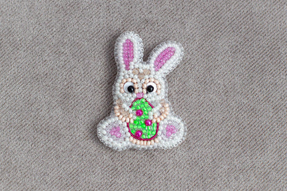A beaded Easter bunny with a green egg in its mouth, sitting on a textured gray background.