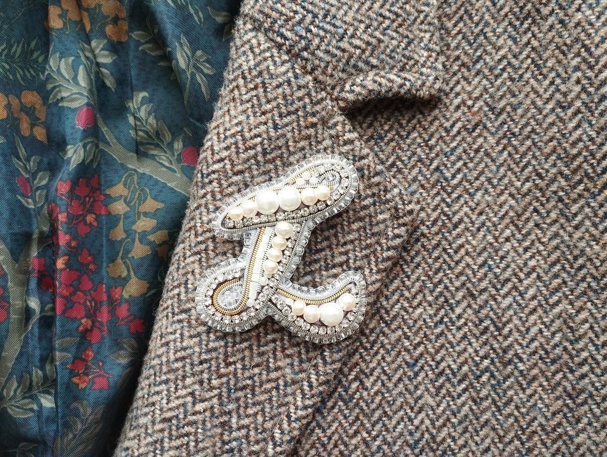 A close-up image of a decorative monogram brooch on a tweed jacket, with a floral patterned fabric visible in the background.