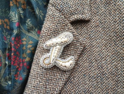 A close-up image of a decorative monogram brooch on a tweed jacket, with a floral patterned fabric visible in the background.