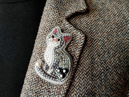 A close-up image of a decorative cat brooch or pin, made of beads and pearls, attached to a tweed jacket or blazer.