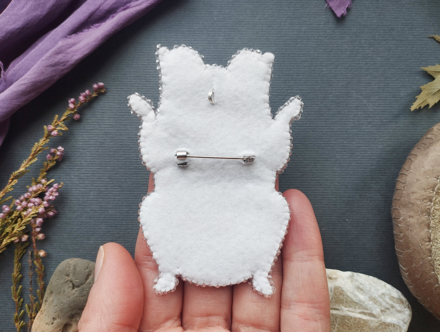 A hand holding a white felt animal pin against a dark background with some purple flowers and a brown object.