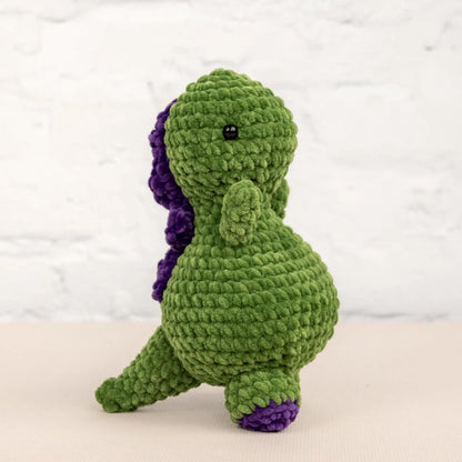 A green crocheted dinosaur toy with purple accents is sitting on a white surface against a white background.