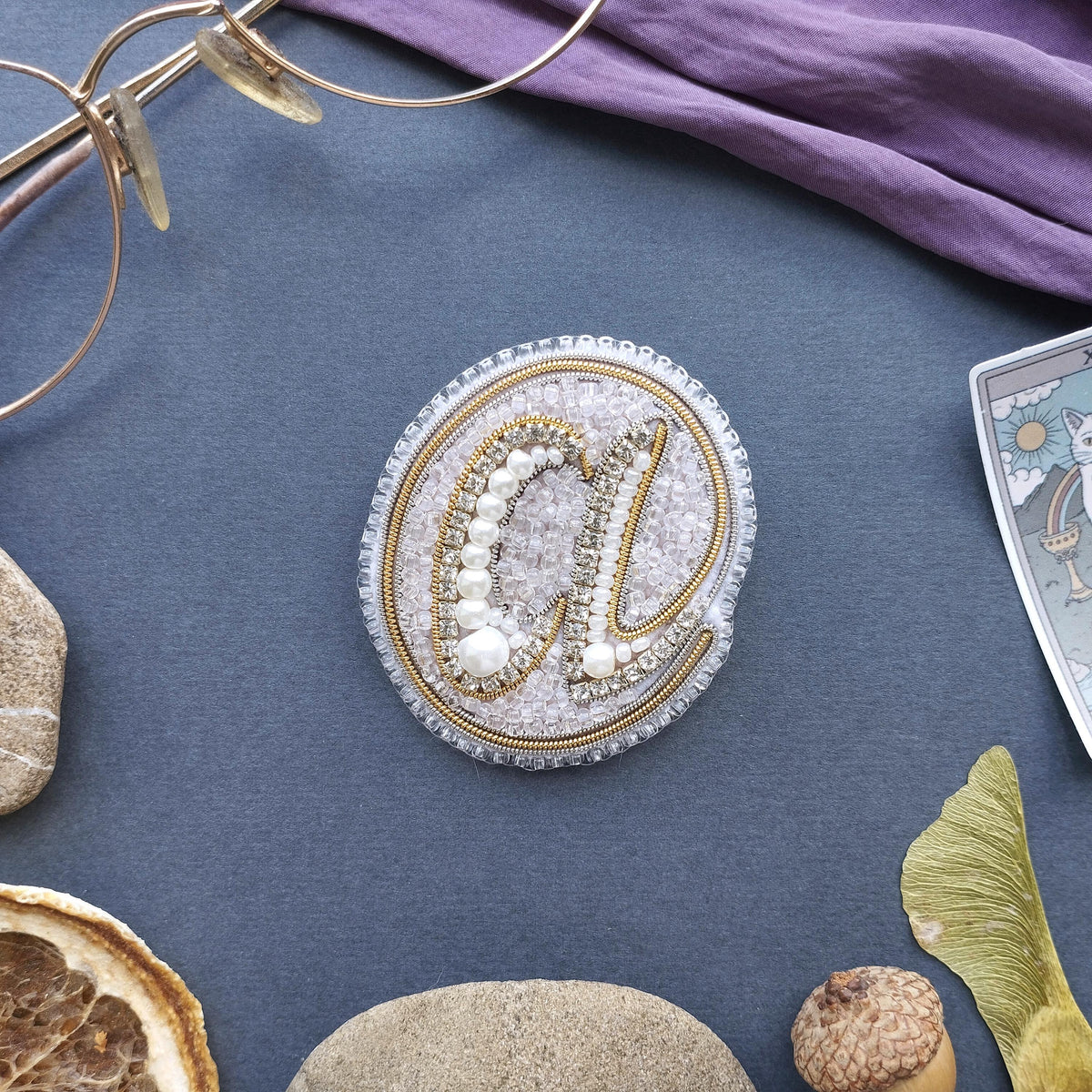 a close-up view of a decorative piece with a letter "a" made of beads and pearls, surrounded by various objects such as a pair of glasses, a purple fabric, and some natural elements like a leaf and a rock.
