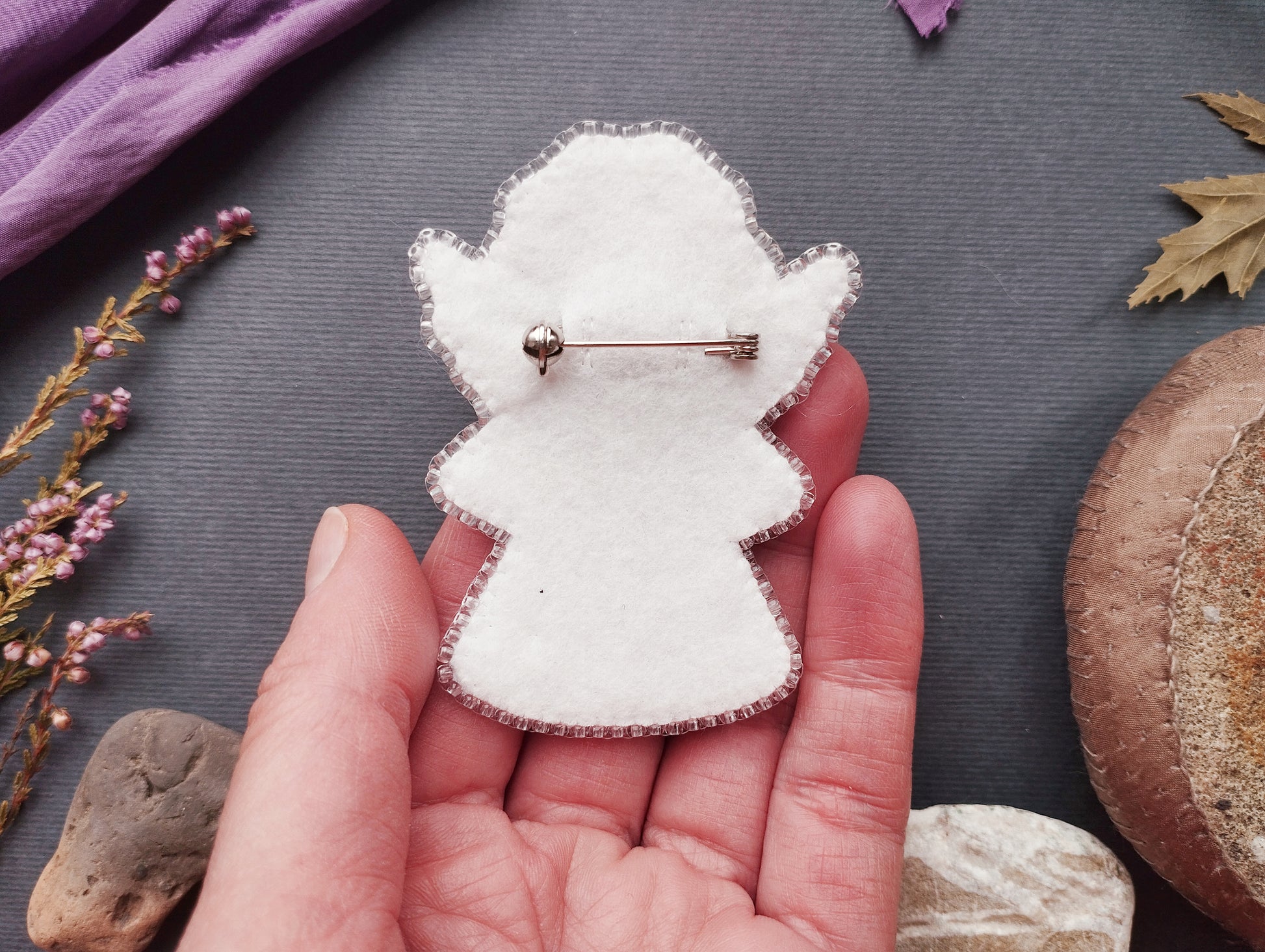 A hand holding a white felt pin or badge against a dark background with various objects, including flowers, rocks, and leaves.