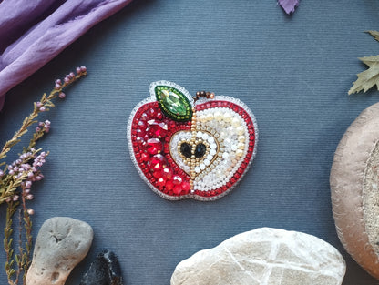 a decorative piece of jewelry, specifically a brooch, with a red and white apple design. The brooch is adorned with various gemstones and pearls, giving it a vibrant and elegant appearance. The piece is placed on a gray surface, surrounded by other objects such as rocks, leaves, and a purple fabric.