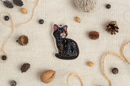 a black cat embroidered on a white background, surrounded by various natural elements such as pine cones, acorns, and a string of beads.