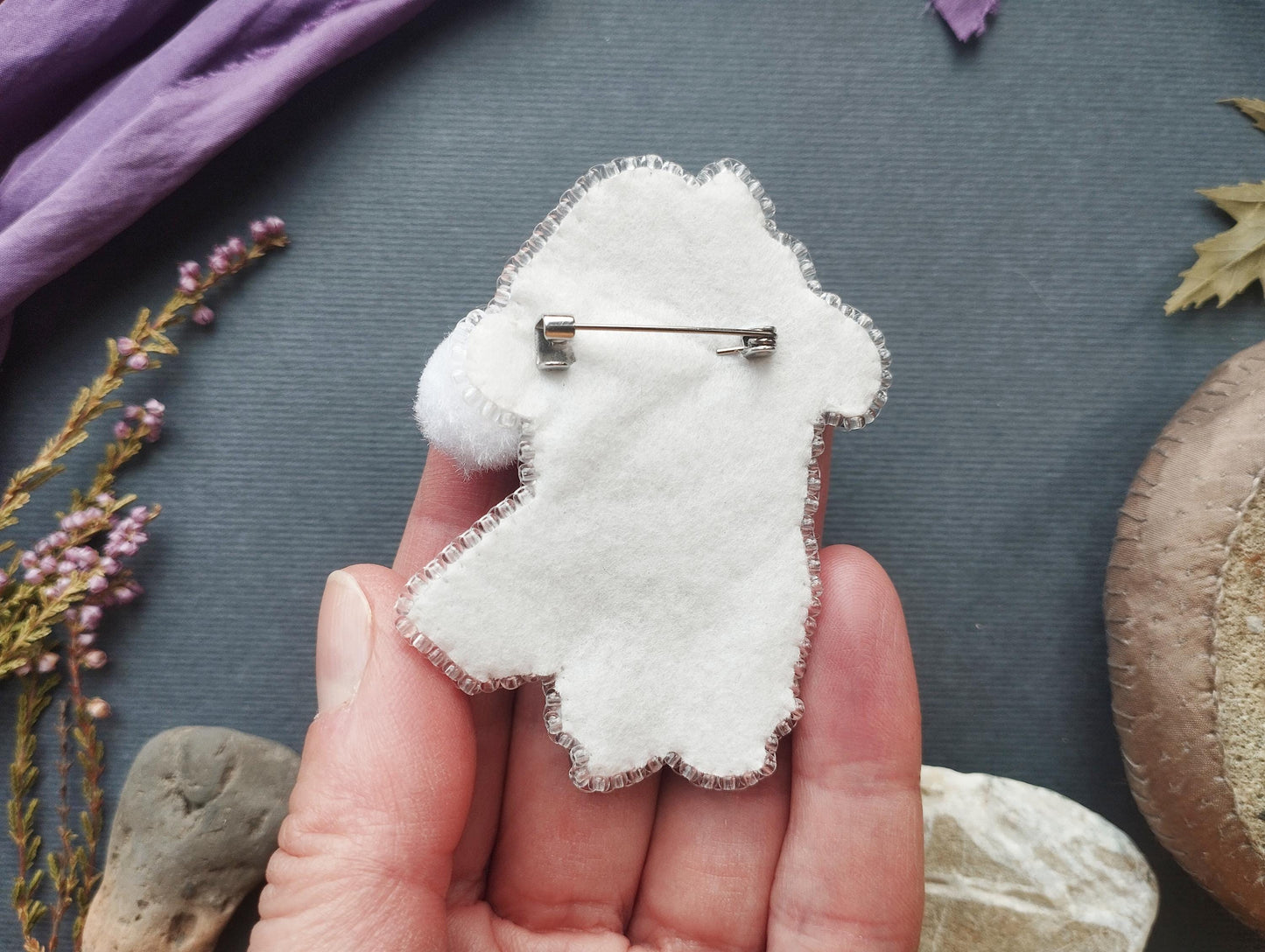A hand holding a white felt animal pin against a dark background with various natural elements, such as flowers and rocks.