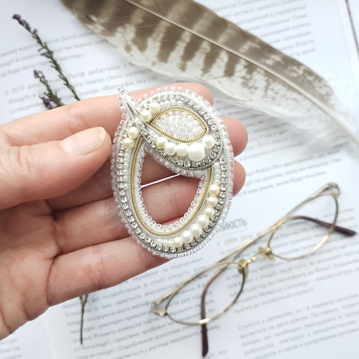 A hand holding a decorative brooch with pearls and rhinestones, alongside a pair of glasses and a feather.