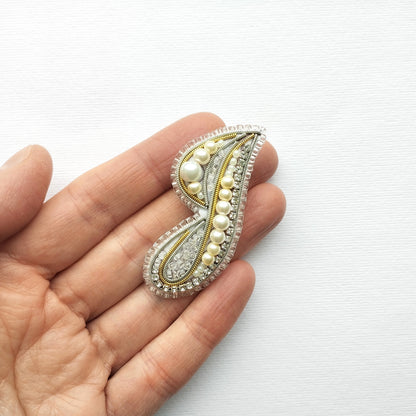 A hand holding a decorative piece of jewelry, which appears to be a brooch or pin, against a white background.