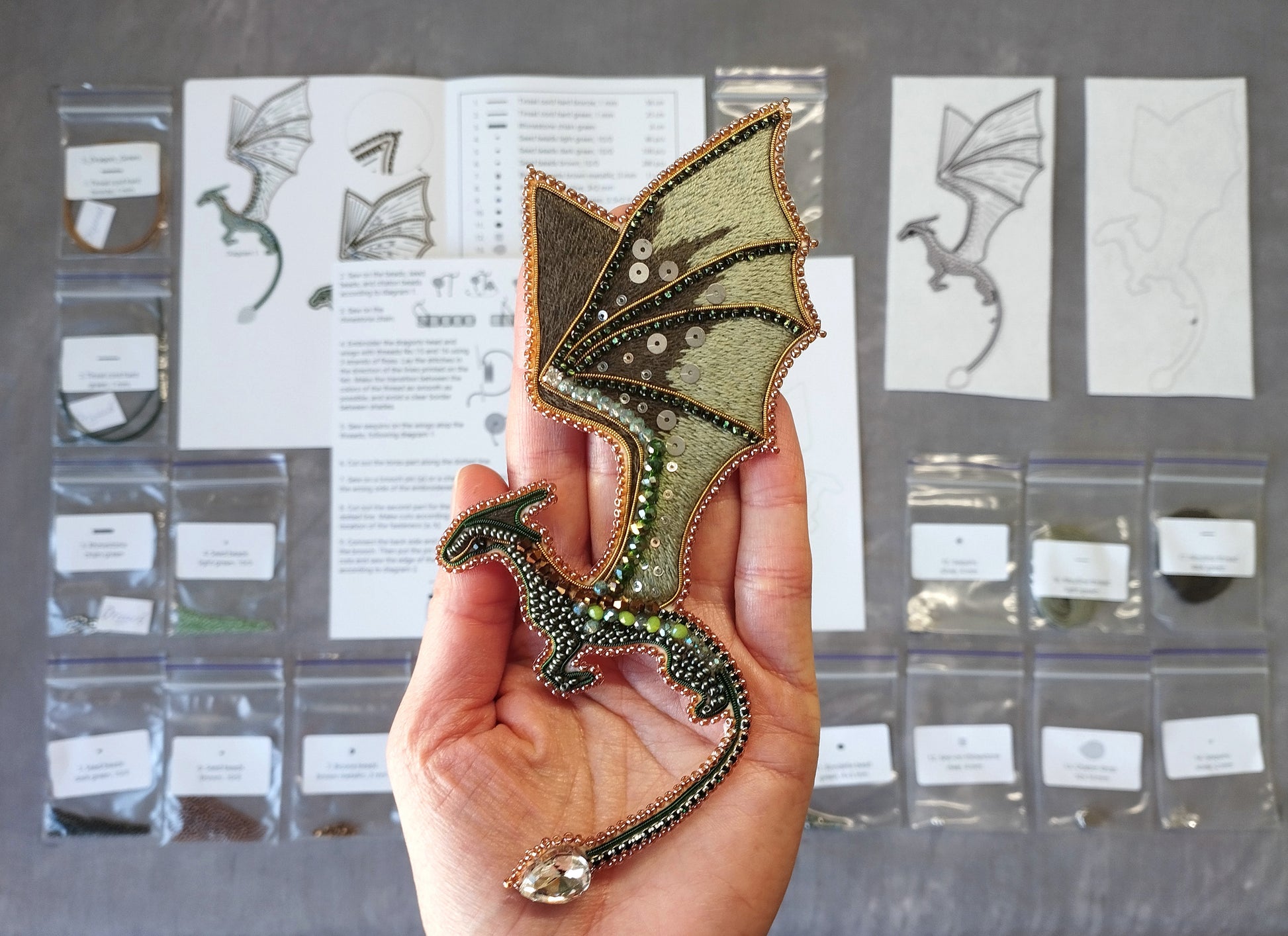 A hand holding a decorative dragon pin made of beads and wire, with various craft supplies and materials visible in the background.