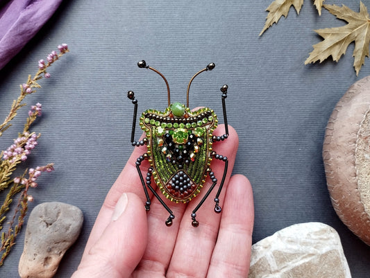 A hand holding a green and black beaded insect brooch against a gray background with various natural elements such as leaves and rocks.