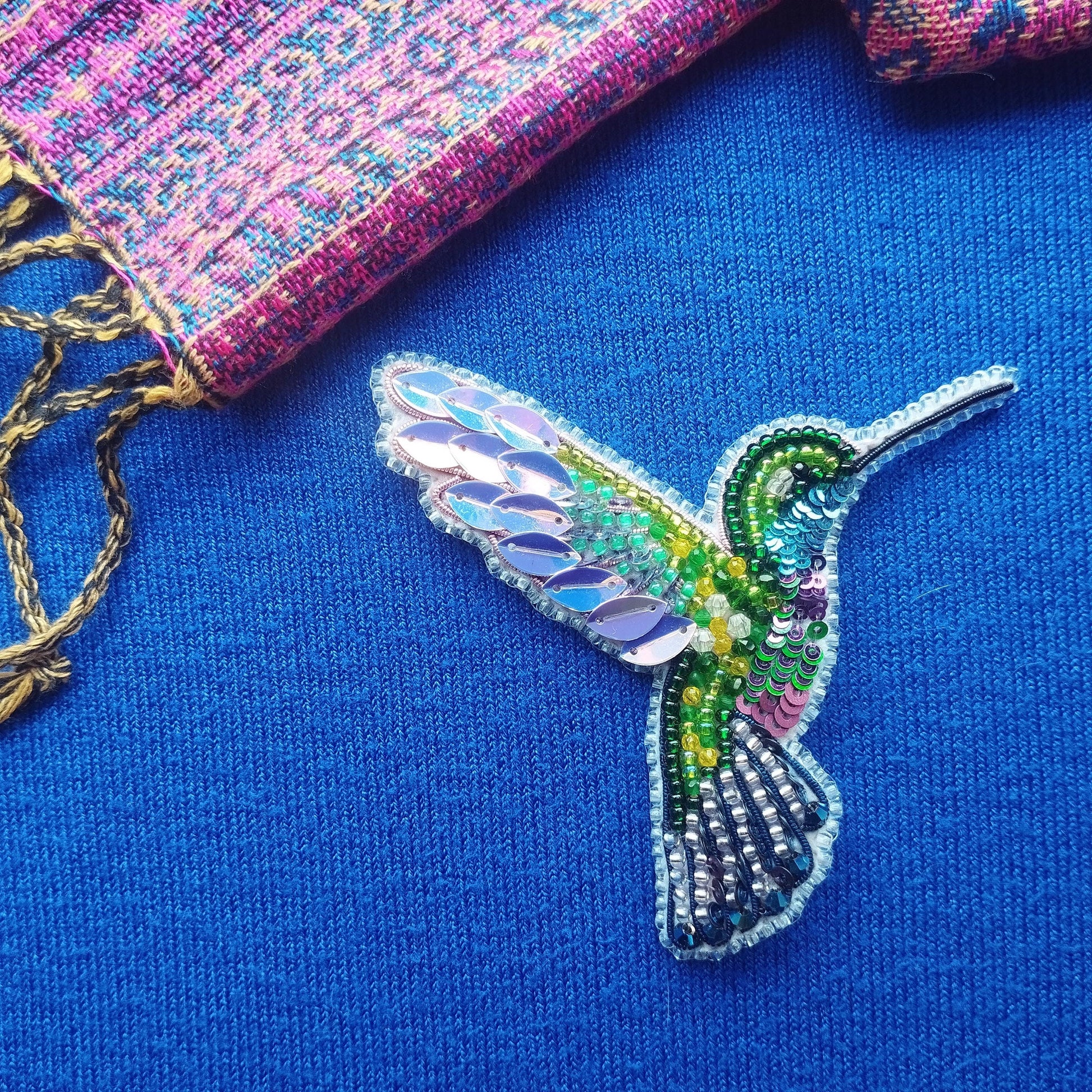 A colorful, intricately designed hummingbird brooch made of beads and sequins is displayed against a blue background.