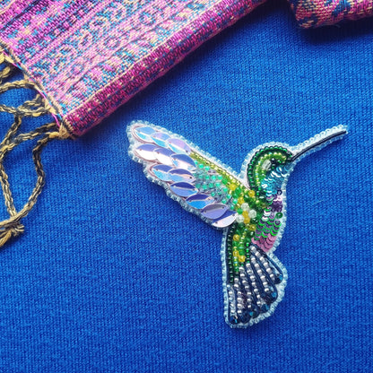 A colorful, intricately designed hummingbird brooch made of beads and sequins is displayed against a blue background.