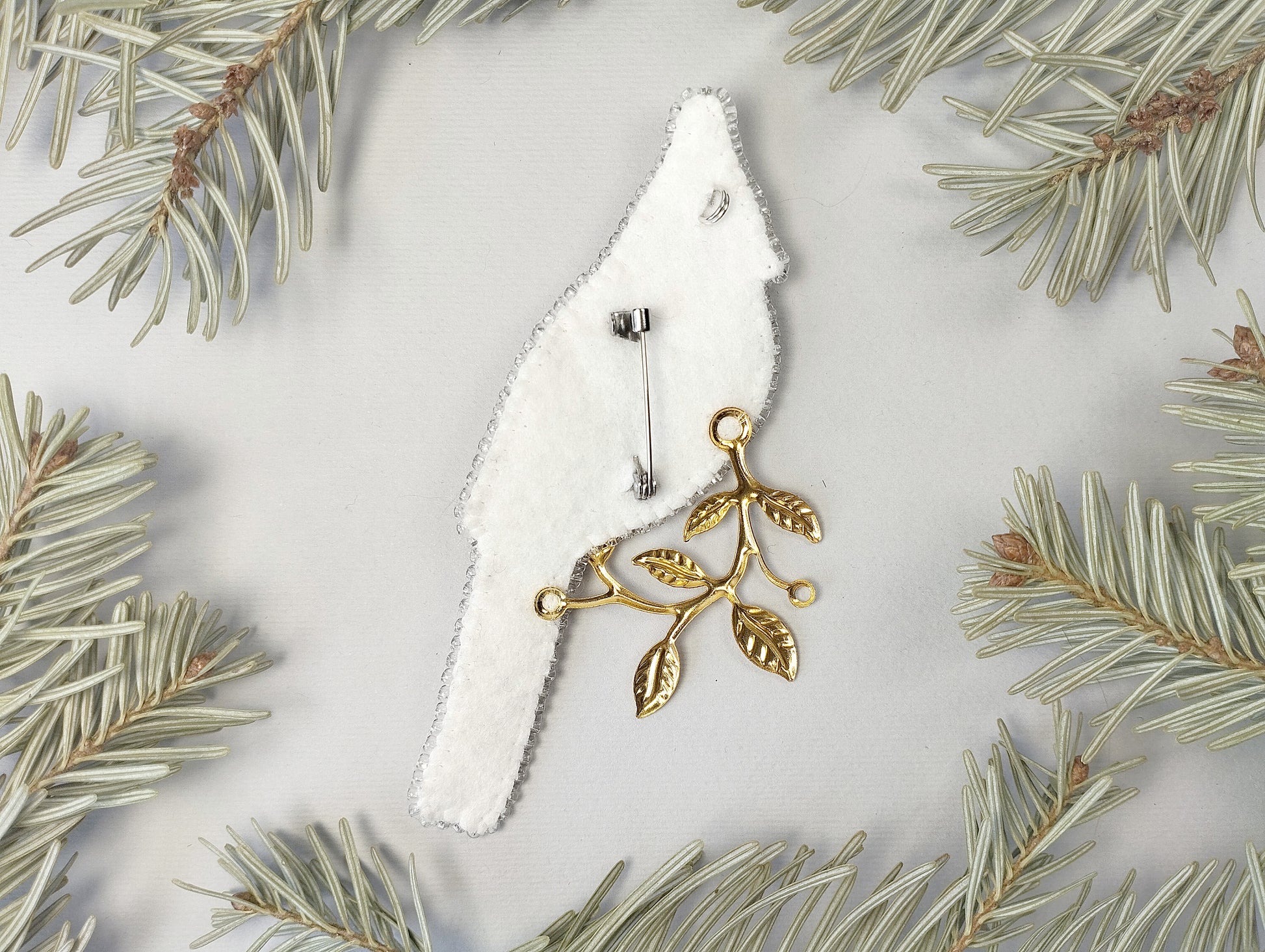 a white bird-shaped pin with a gold leaf design, surrounded by pine needles on a light background.