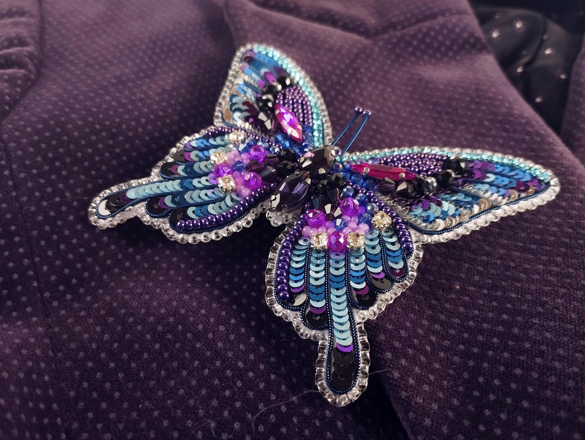 A colorful, intricately designed butterfly brooch made of beads and sequins, resting on a purple fabric.