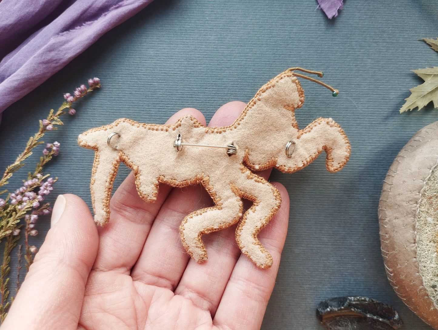 A hand holding a handmade embroidered or sewn object resembling a horse or mythical creature, with some purple flowers and a brown object in the background.