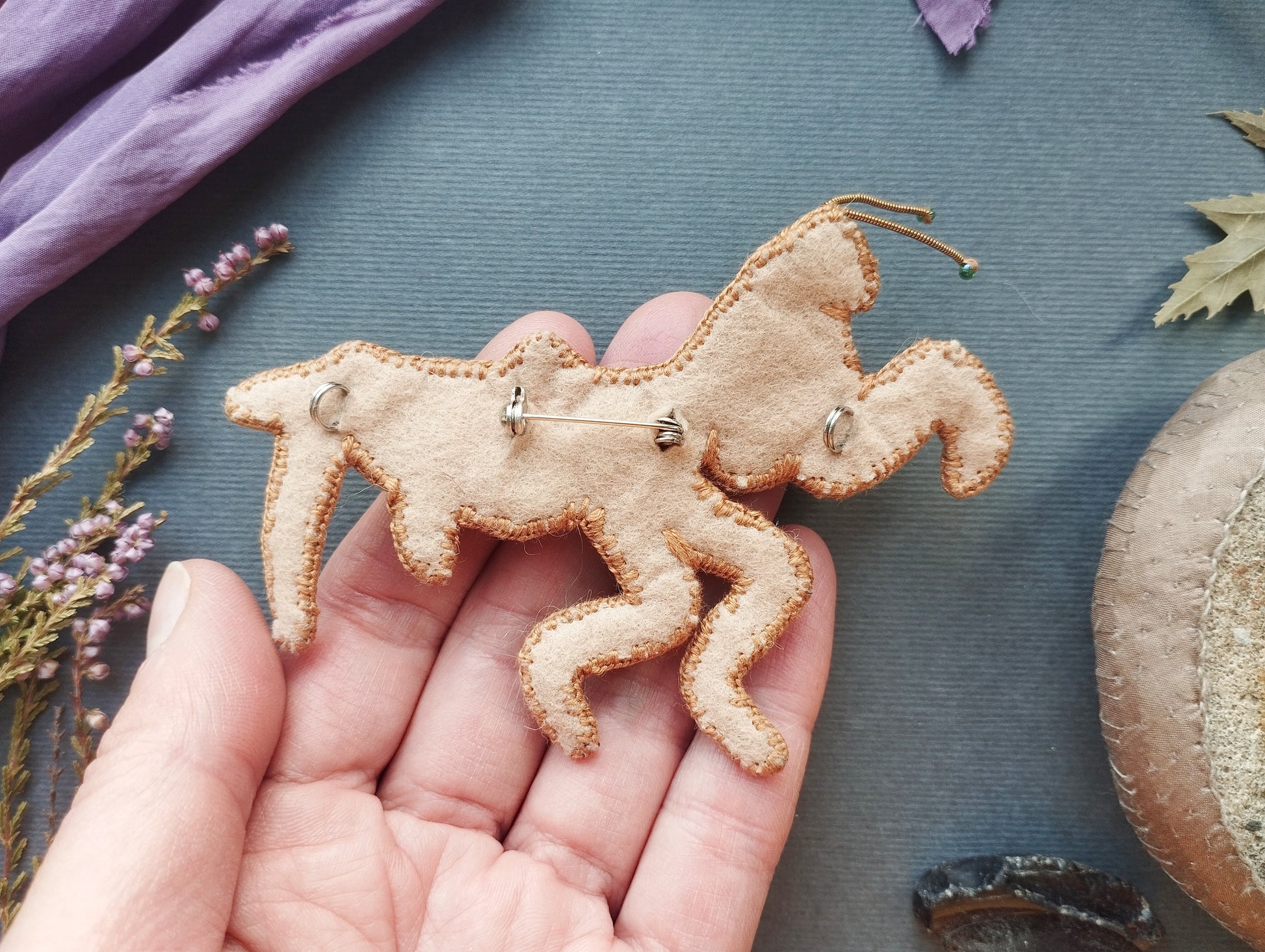 A hand holding a handmade embroidered or sewn object resembling a horse or mythical creature, with some purple flowers and a brown object in the background.