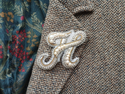 A close-up image of a decorative brooch or pin with pearls and a metallic design, attached to a tweed jacket or blazer.