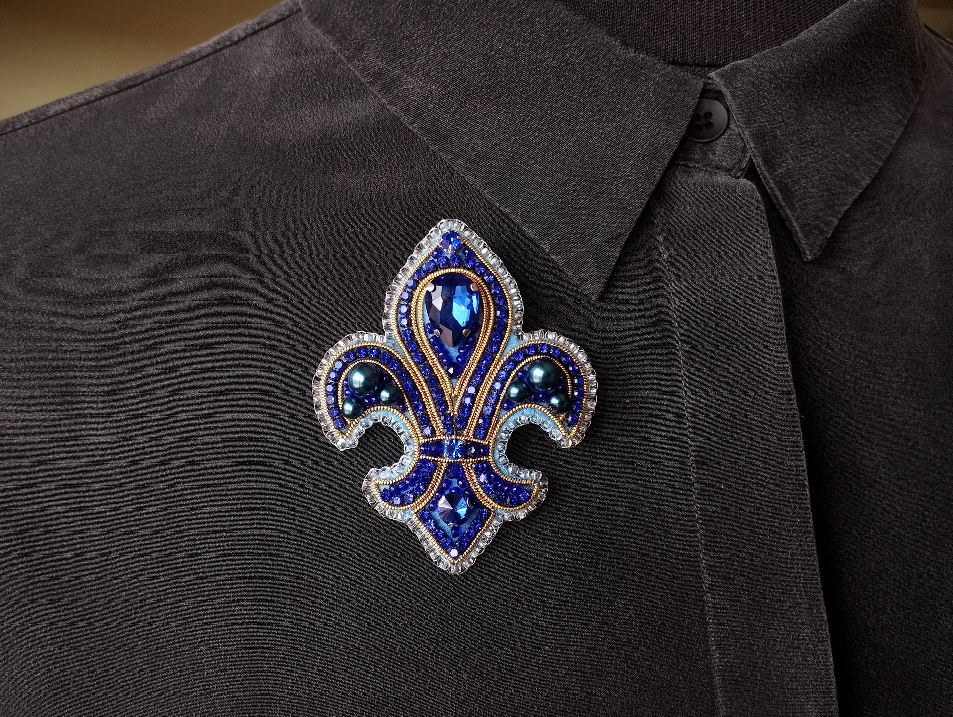 A close-up image of a decorative brooch on a dark fabric, featuring a blue and gold design with a central blue gemstone surrounded by smaller blue gemstones.