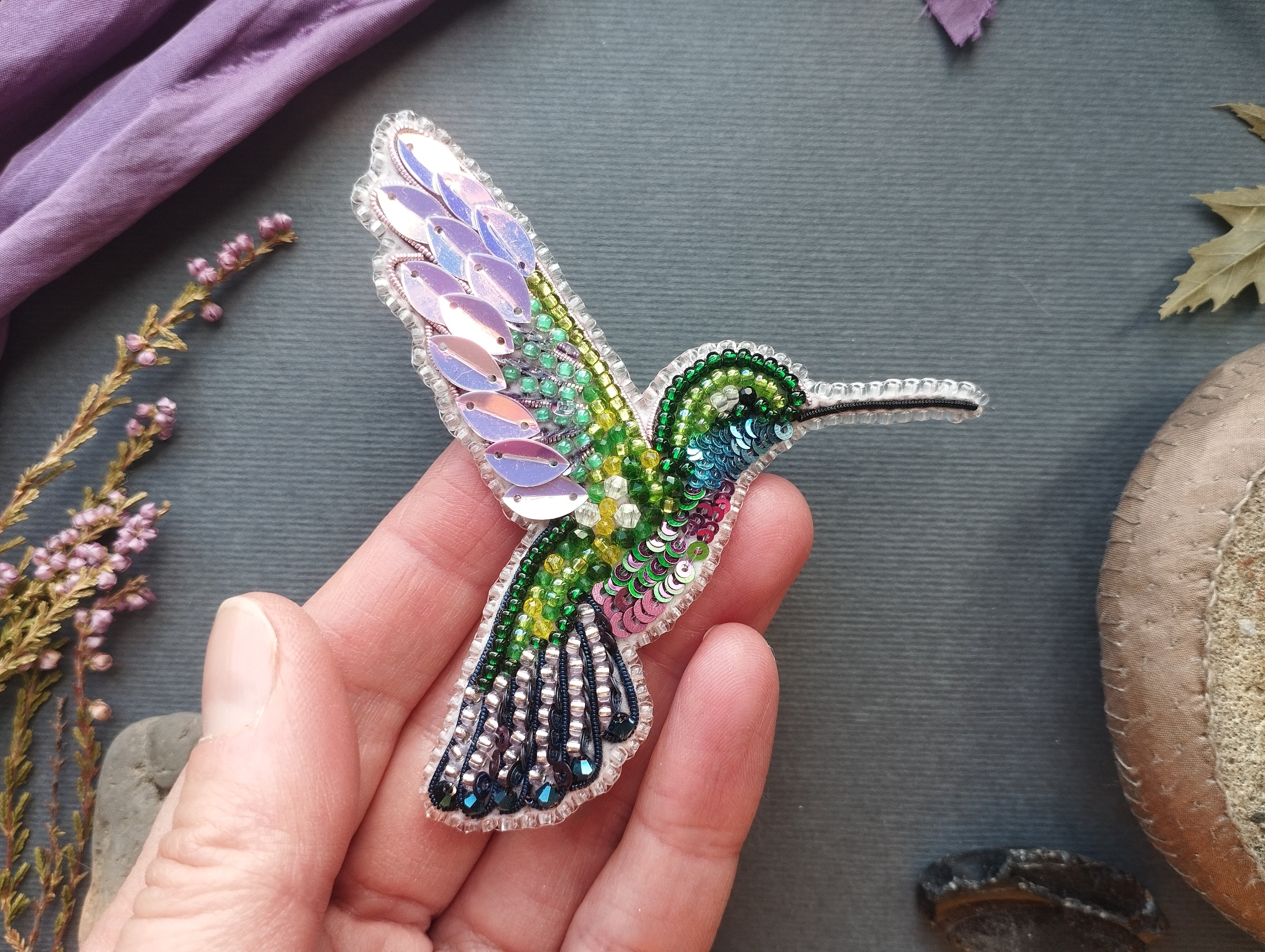 A hand holding a colorful, intricately beaded hummingbird brooch against a gray background.