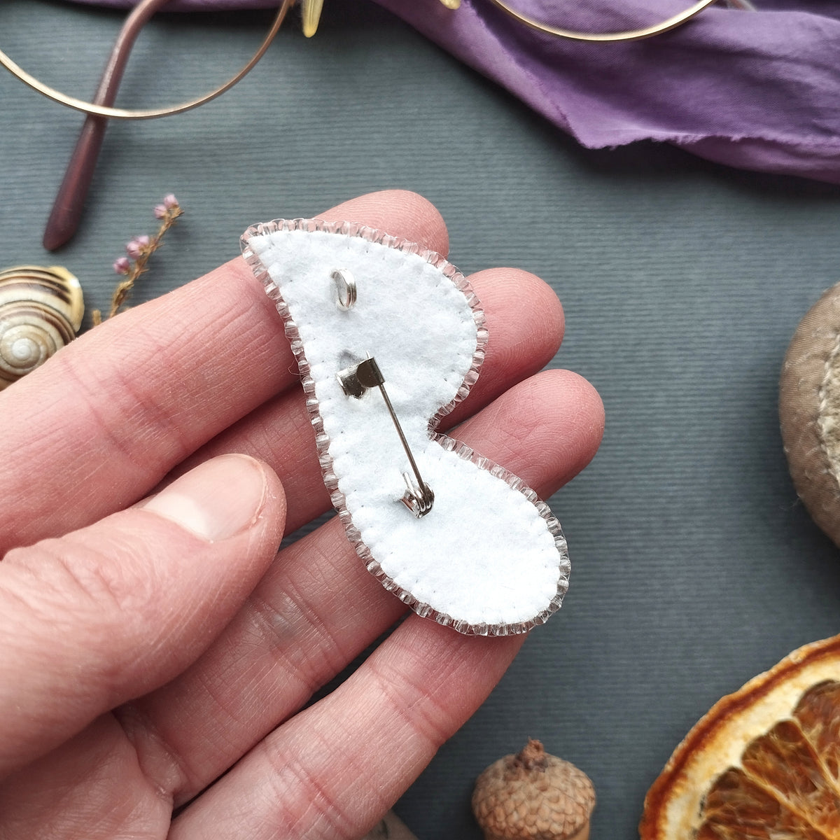 A hand holding a white felt pin with a silver pin attached to it, surrounded by various objects such as a pair of glasses, a purple fabric, and dried fruit.