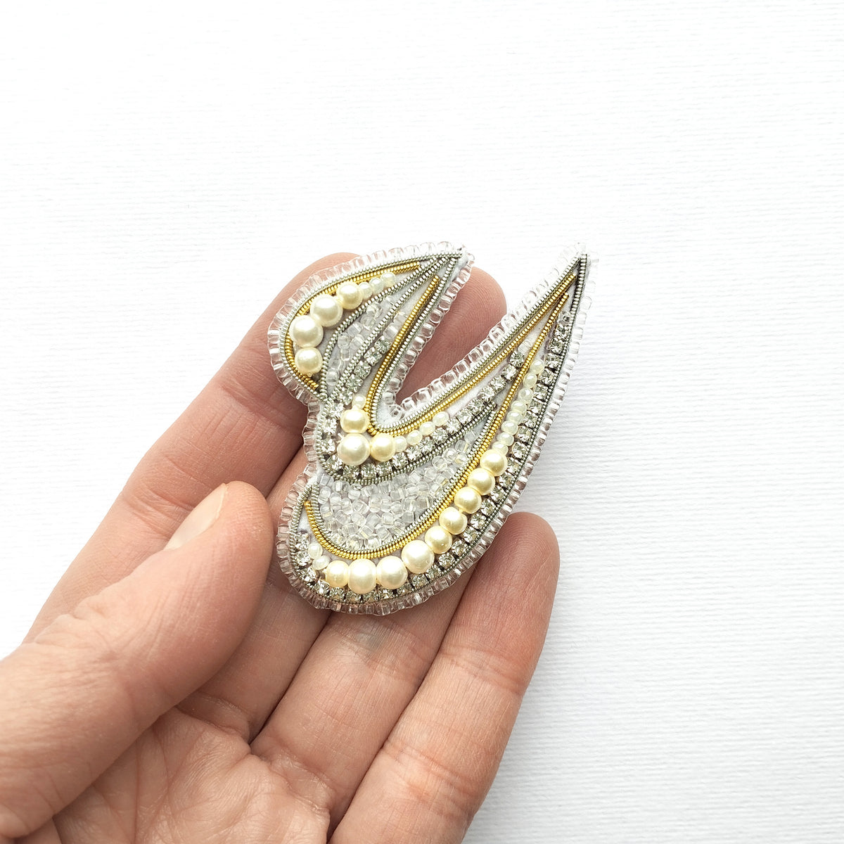 A hand holding a decorative piece of jewelry, which appears to be a brooch or pin, against a white background.