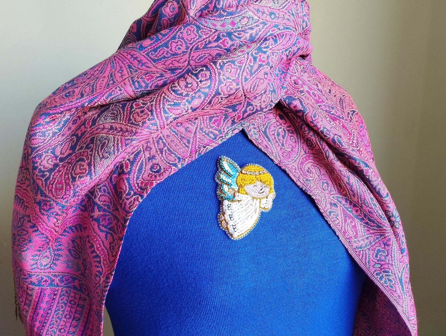 A close-up of a person wearing a blue shirt with a colorful, patterned scarf draped over it. The scarf has a paisley design in shades of pink and purple. There is a small, embroidered pin or brooch on the person's shirt, featuring an angel-like figure.