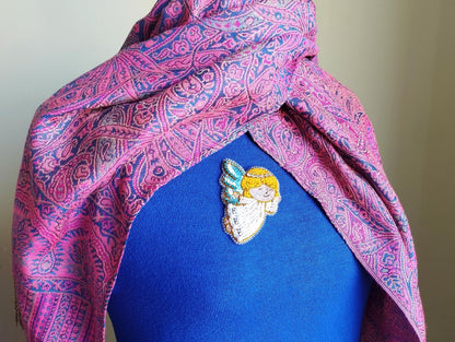 A close-up of a person wearing a blue shirt with a colorful, patterned scarf draped over it. The scarf has a paisley design in shades of pink and purple. There is a small, embroidered pin or brooch on the person's shirt, featuring an angel-like figure.