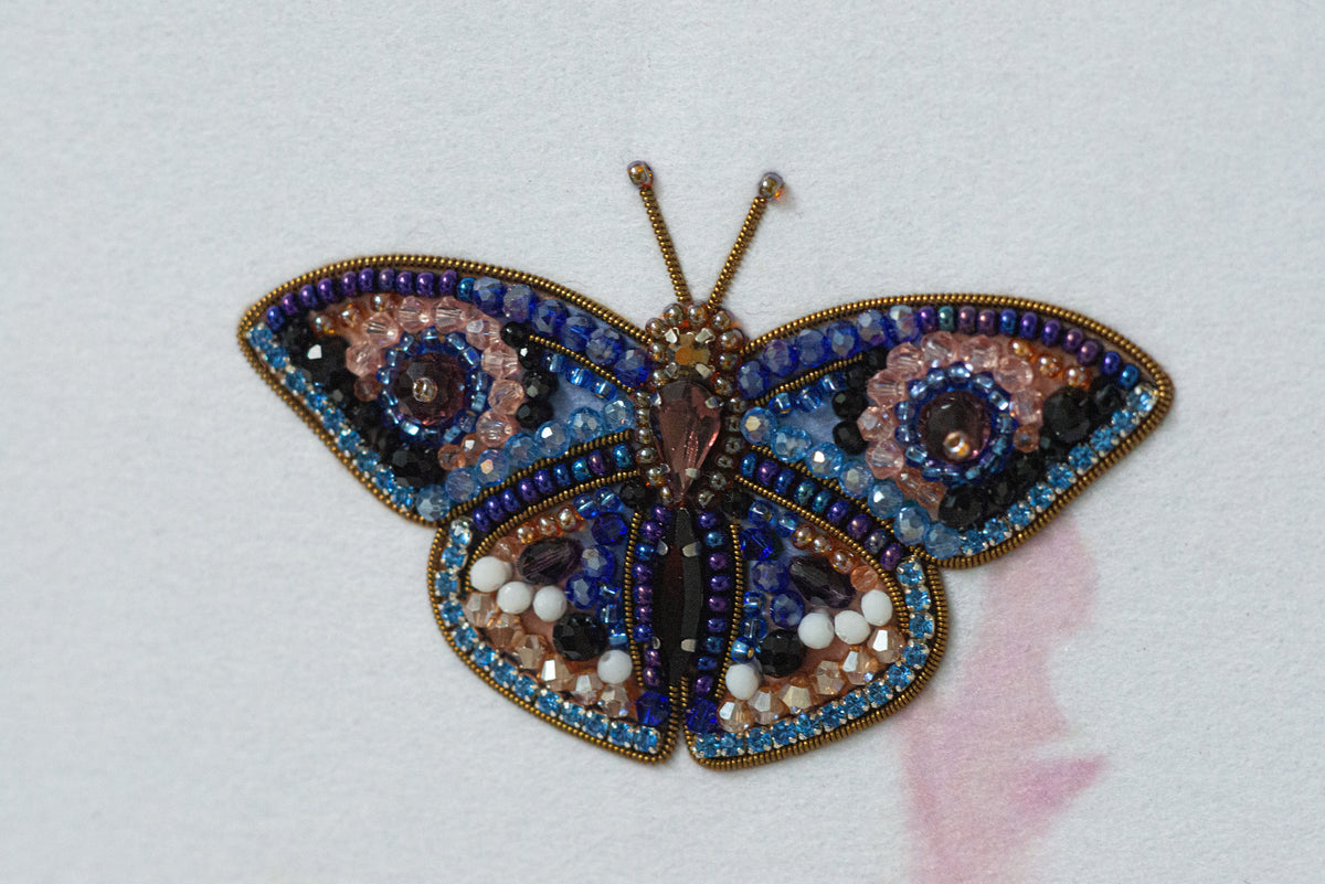 A colorful, intricately designed butterfly brooch made of various materials, including beads, stones, and sequins, is displayed against a white background.