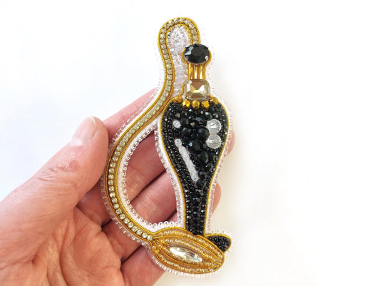 A hand holding a decorative piece of jewelry, which appears to be a pendant or brooch. The piece is adorned with a combination of black and gold elements, including a central black figure surrounded by a gold chain and embellished with rhinestones and crystals.