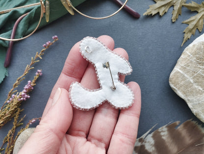 A hand holding a white felt object with a pin hole in the center, surrounded by various natural elements such as leaves, feathers, and flowers.