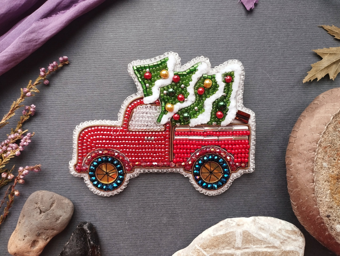 A red and white embroidered Christmas tree ornament in the back of a red pickup truck, surrounded by various natural elements such as leaves, rocks, and flowers.