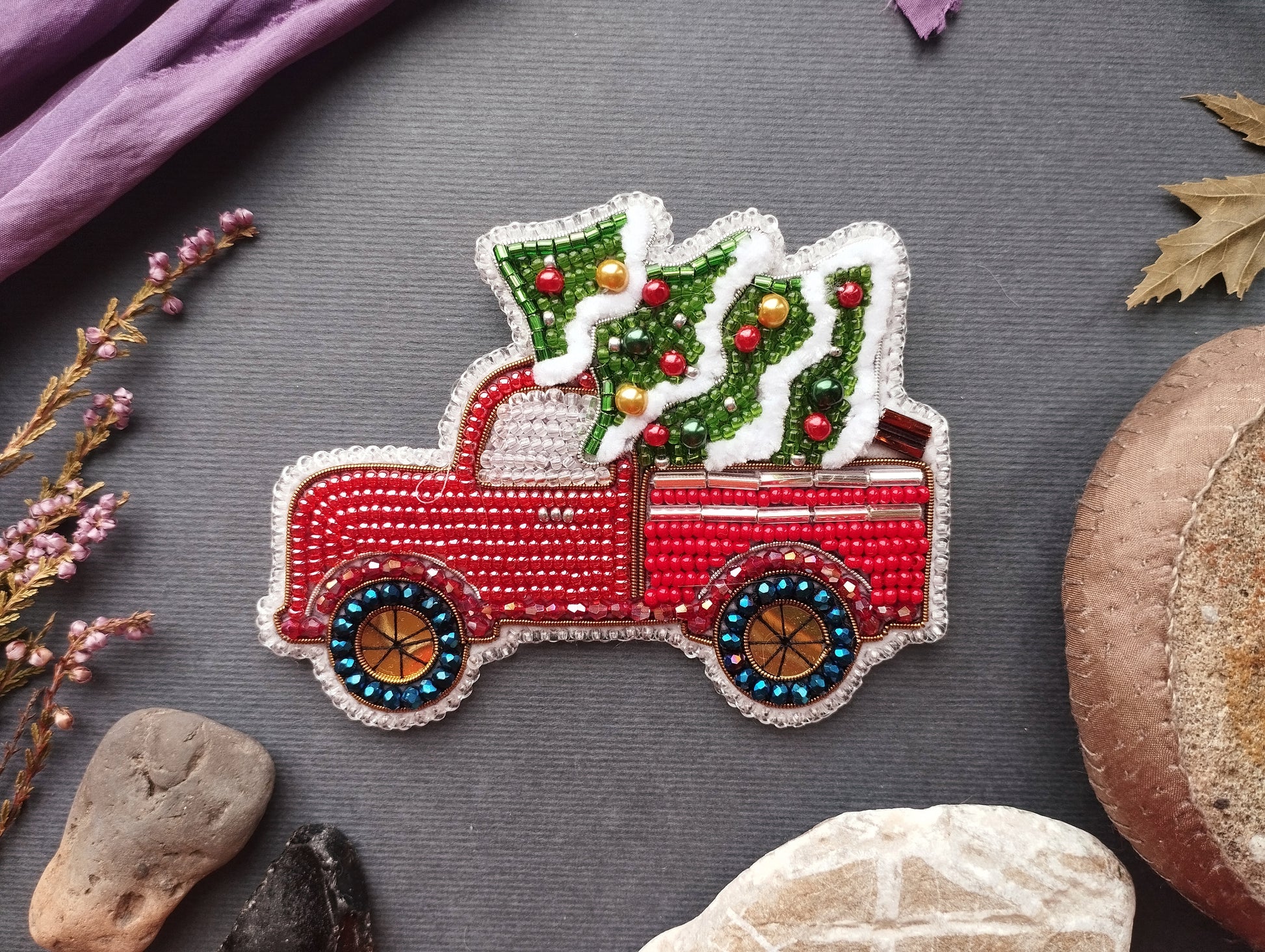 A red and white embroidered Christmas tree ornament in the back of a red pickup truck, surrounded by various natural elements such as leaves, rocks, and flowers.