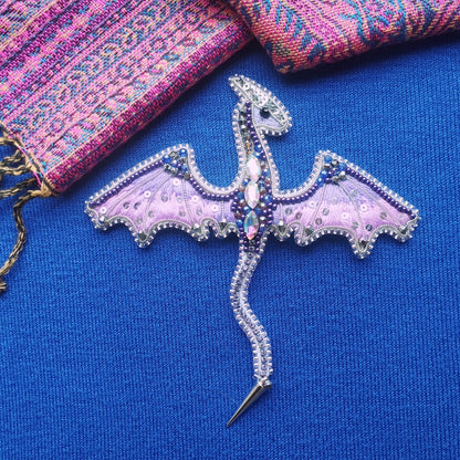 A decorative dragon brooch with intricate beading and gemstone details is displayed against a blue background.