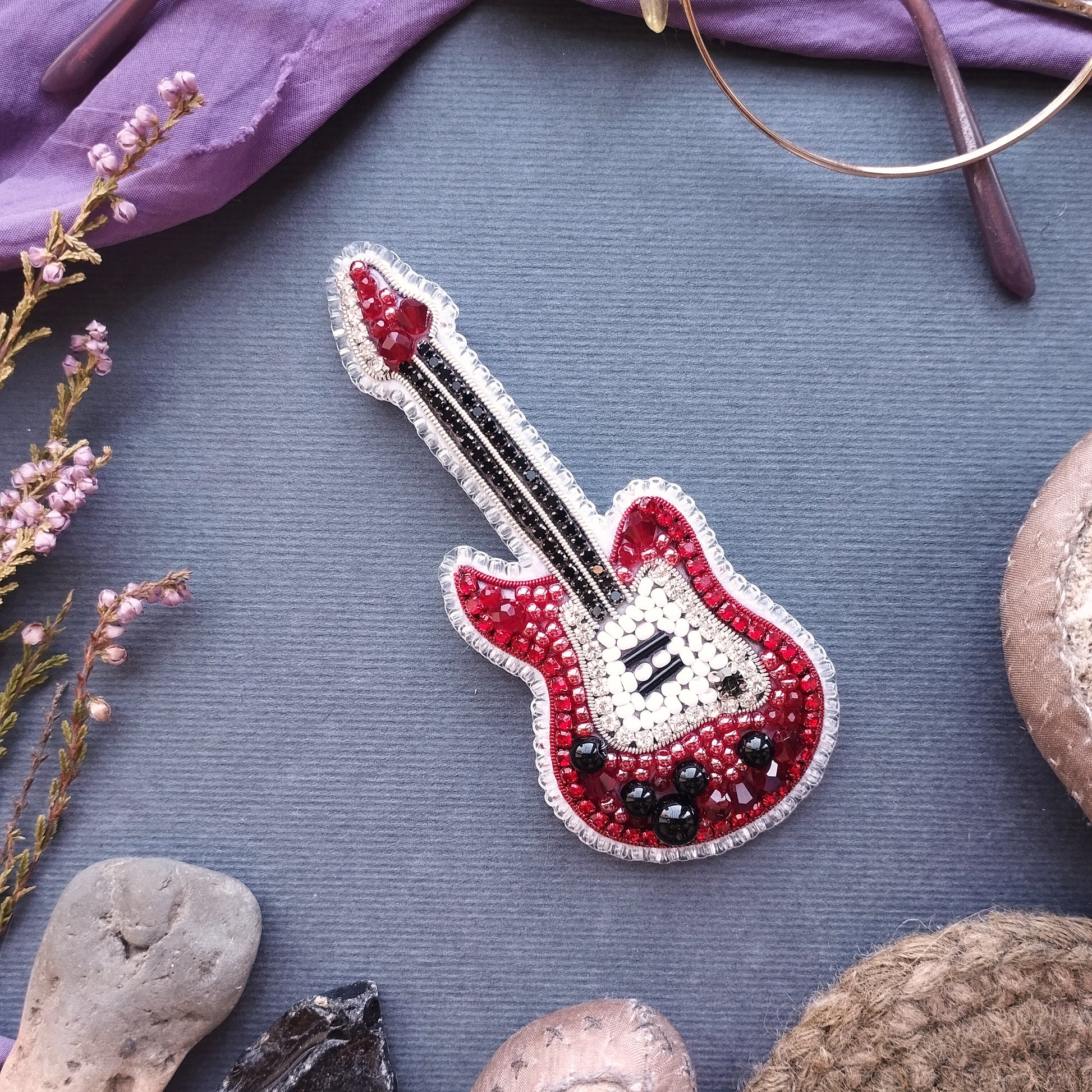 a red and white guitar brooch with a beaded design, placed on a gray surface surrounded by various objects such as a pair of glasses, a plant, and a rock.