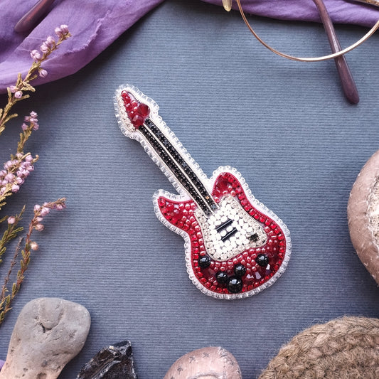 a red and white guitar brooch with a beaded design, placed on a gray surface surrounded by various objects such as a pair of glasses, a plant, and a rock.