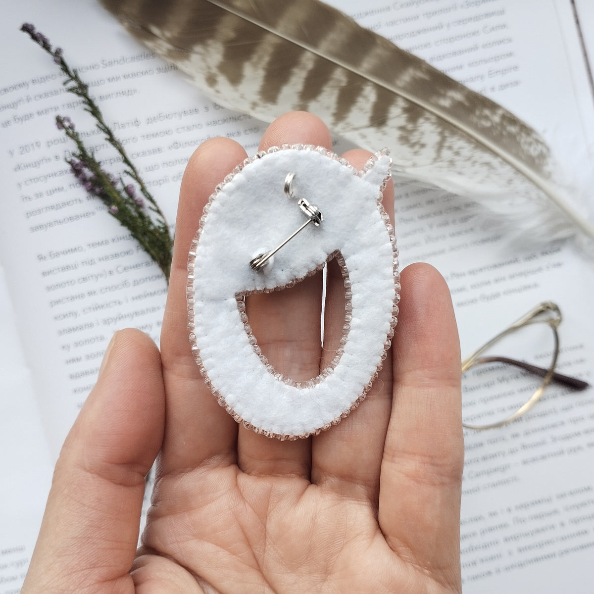 A hand holding a white fabric pin with a smiley face design, surrounded by feathers and a pair of glasses on a page of text.
