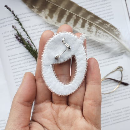 A hand holding a white fabric pin with a smiley face design, surrounded by feathers and a pair of glasses on a page of text.