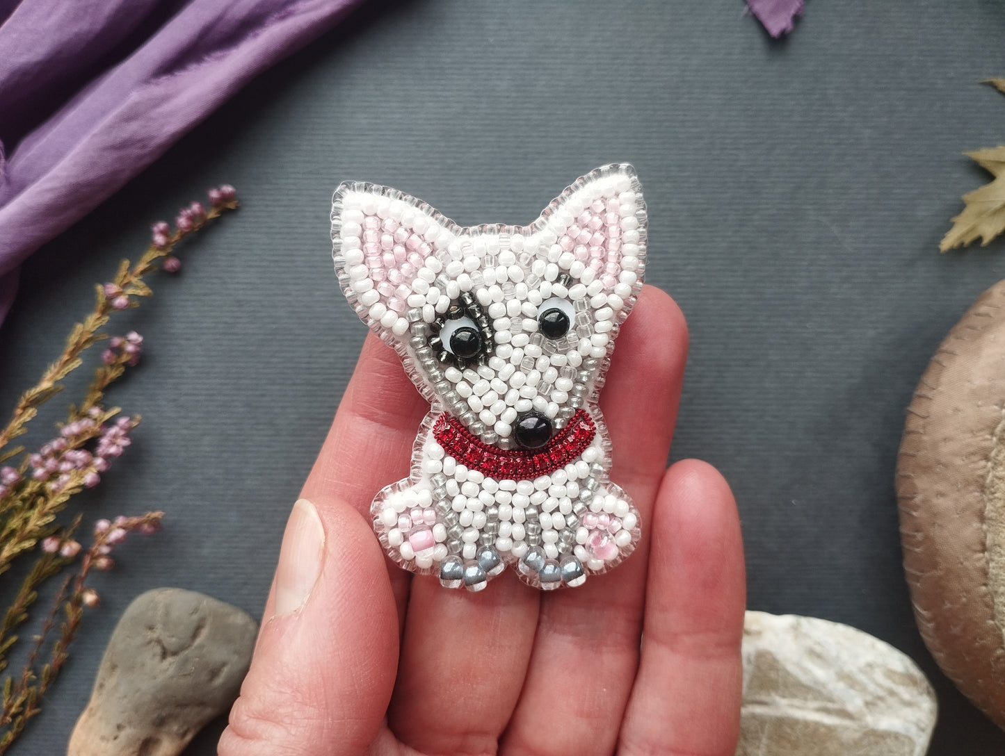 A hand holding a small, intricately beaded embroidered dog-shaped brooch against a dark background.