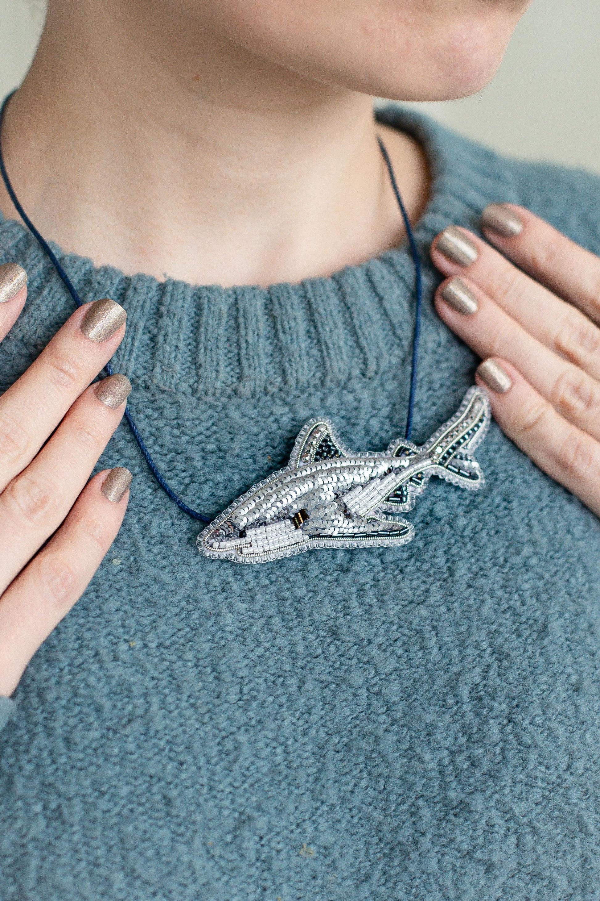A close-up of a person's neck and chest wearing a blue sweater, with their hands placed on their chest. They are wearing a necklace with a silver, intricately designed shark pendant.