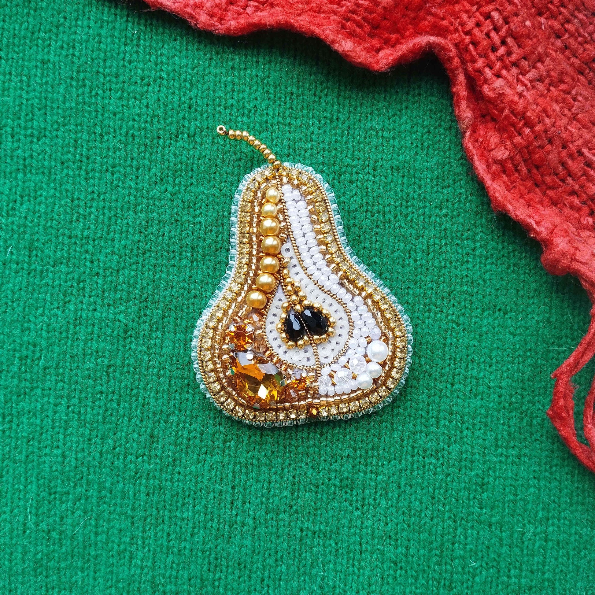 A decorative piece featuring a pear-shaped design with gold and white beads, black stones, and a red fabric on a green background.