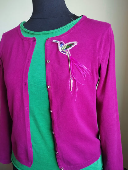 A mannequin wearing a pink cardigan with a green shirt underneath, and a colorful bird brooch on the front of the cardigan.