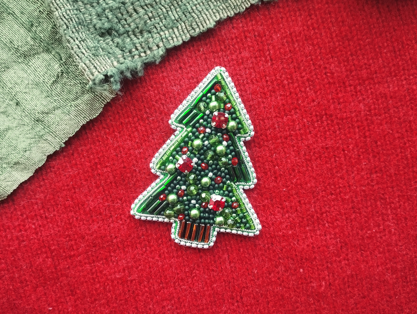 A green and red beaded Christmas tree brooch is displayed on a red fabric surface.