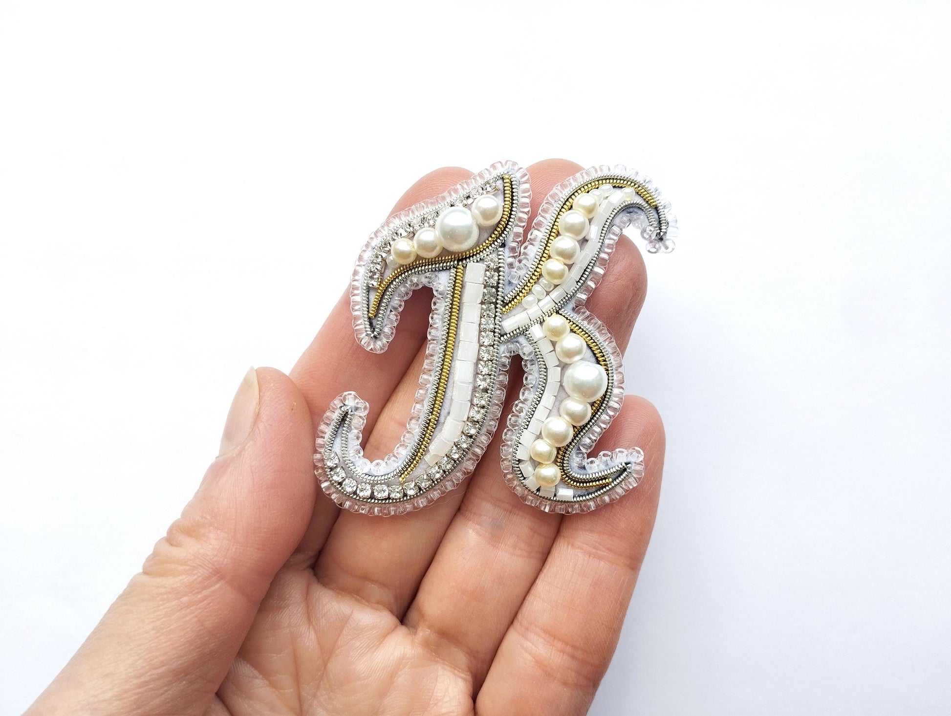A hand holding a decorative letter 'K' made of beads, pearls, and rhinestones.
