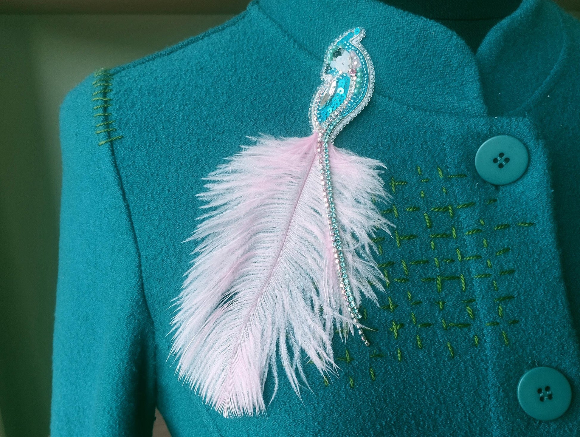 A close-up of a teal-colored jacket with a feather pin and buttons on the front.