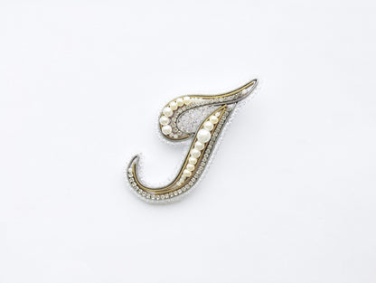 A decorative letter 'J' made of various materials, including pearls and diamonds, against a white background.