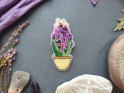 a decorative piece of embroidered art, which appears to be a potted plant or flower, placed on a grey background. Surrounding the embroidered piece are various natural elements, including a purple flower, a leaf, and some rocks.