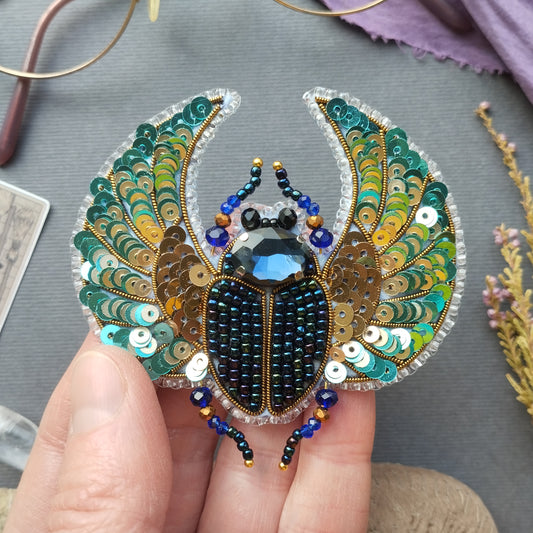 A hand is holding a decorative piece of jewelry featuring a detailed embroidered design of a colorful beetle.
