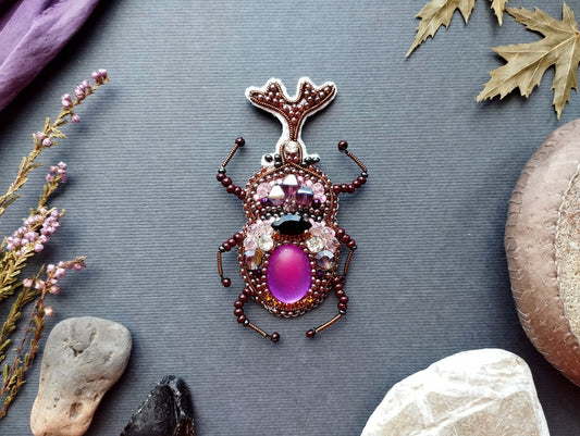 a detailed embroidered insect brooch with a purple center and various embellishments, including gemstones and beads, set against a gray background.