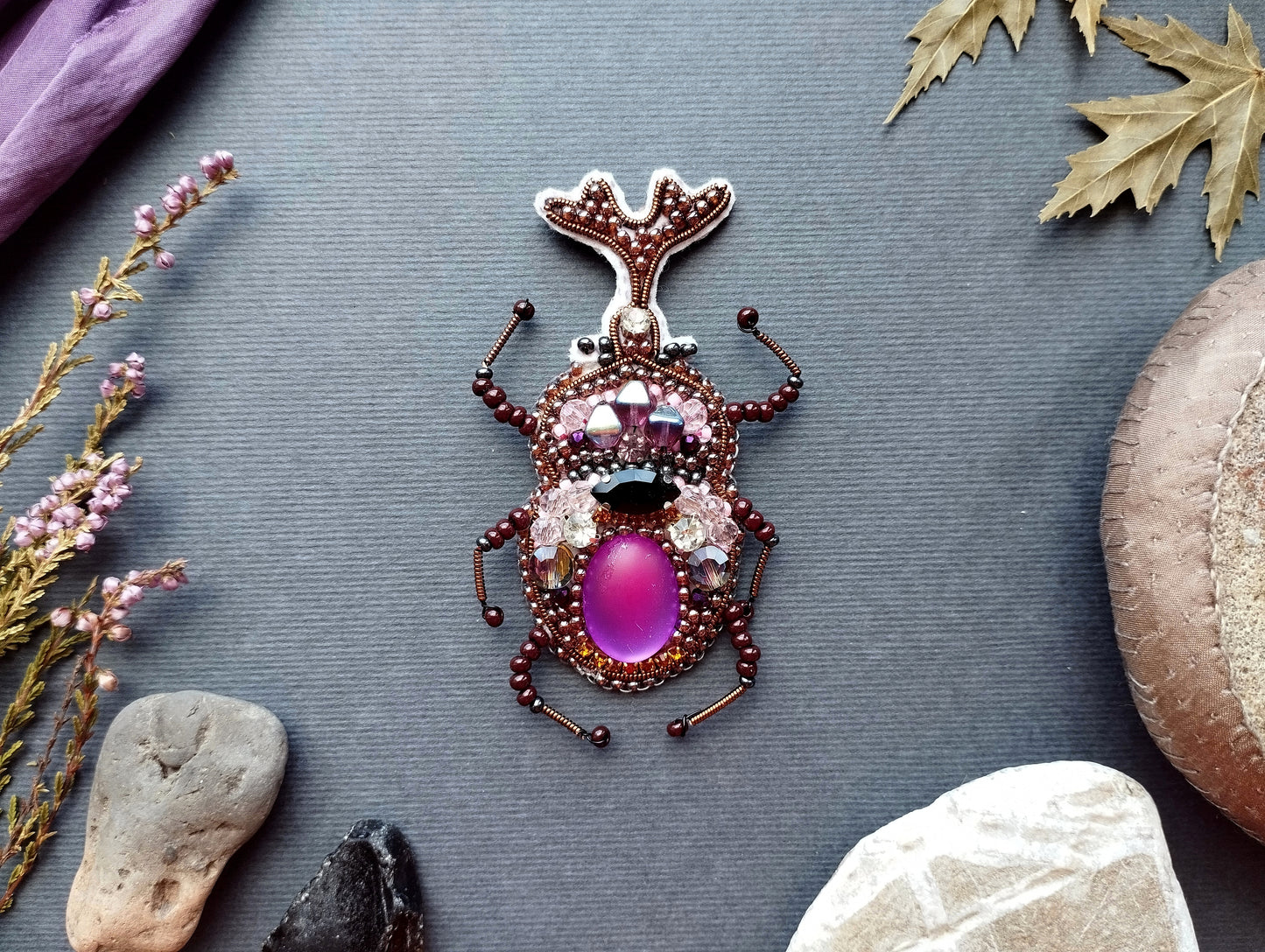 a detailed embroidered insect brooch with a purple center and various embellishments, including gemstones and beads, set against a gray background.
